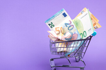 roll of euro banknotes with white ribbon in supermarket trolley cart isolated on purple background