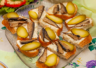 Fototapeta na wymiar Glass plate with sandwiches with smoked sprat and pickles close-up on the background of a colored tablecloth table