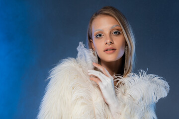 young pierced woman in white faux fur jacket with feathers posing on dark blue background.