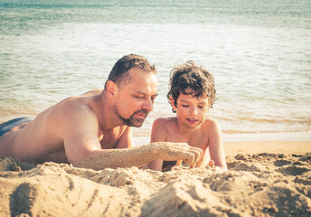 Happy mature father and son at sea coast. Mid adult man and schoolboy lying on the sand. Family, vacation, outdoor activity concept