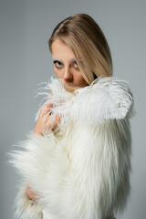 young woman in white faux fur jacket holding feather and looking at camera isolated on grey.