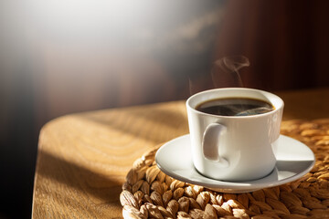 Coffee cup with hot steam on brown wood table with morning sunlight shining through the window. Healthy black coffee in a mug. Copy space is on the left side. Light and shadow concept. - obrazy, fototapety, plakaty
