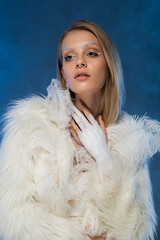 pierced young woman with winter makeup posing in white faux fur jacket with feathers on dark blue.