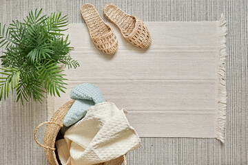Natural muslin home towels, white and soft blue, lie in a wicker basket against a white cotton rug and home wicker slippers. View from above. 