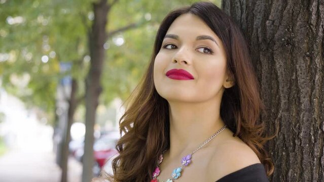 A young beautiful Caucasian woman thinks about something with a smile as she leans against a tree in a park on a sunny day - closeup