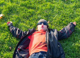 Happy child in spring season lying on green grass. Smiling boy in sunglasses, jacket and hat having fun outdoor in autumn park