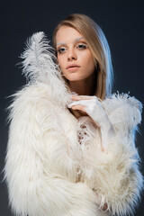 pretty young woman in white faux fur jacket posing with feather isolated on blue.