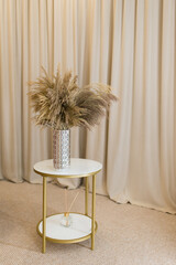 Soft home decor in gentle tones, a tall cylindrical vase with a bouquet of dried flowers against a background of beige curtains, on a round table. Interior.