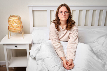 Portrait of an adult woman sitting on a bed with a blanket in a white bedroom. A brunette woman...