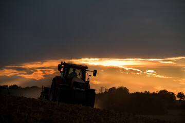 Fototapeta premium Tractor harrowing reddish brown, dusty, clayey soil in a hilly landscape at sunset