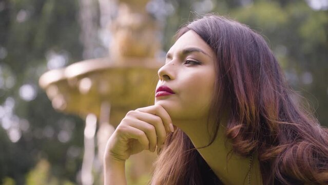 A young beautiful Caucasian woman thinks about something in a garden - closeup - fountain in the blurry background
