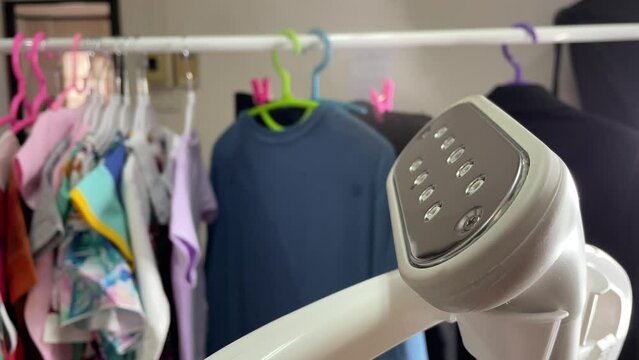 Portable handheld ironing steamer machine with hot steam and cloth on hanger in background. Household appliance concept