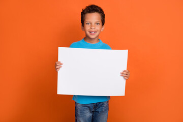 Photo portrait of cute little pupil boy hold empty paper sheet banner poster wear trendy blue outfit isolated on orange color background