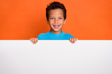 Photo portrait of charming little boy standing behind white banner empty space wear trendy blue clothes isolated on orange color background