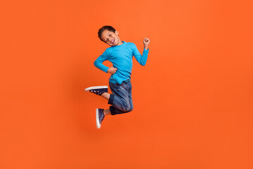 Fototapeta na wymiar Full size photo of charming small pupil boy jump play raise fists celebrate dressed stylish blue look isolated on orange color background