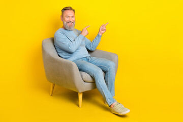 Fototapeta na wymiar Full size portrait of positive aged person sit chair indicate fingers empty space isolated on yellow color background