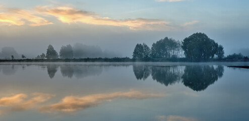 Summer-autumn landscape of trees reflected in the water of the lake on the shore and the sky in the early foggy morning. Panoramic web banner