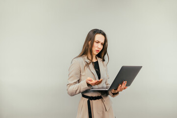 Beautiful female office worker in a jacket stands on a beige background and uses a laptop with a serious face.