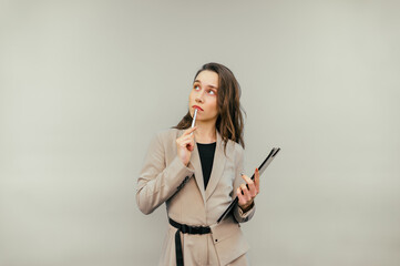 Pensive business woman in a suit stands on a beige background with a paper tablet and a pen in his hands, looks away with a serious face and thinks.