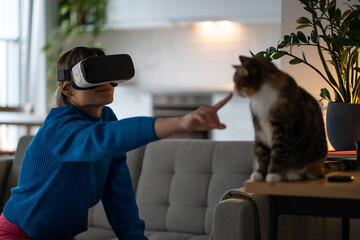Positive casual woman owner in VR headset pulls hand to cat uses AR technology to play with pet....