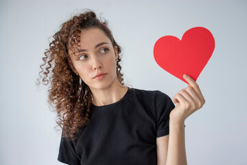 Portrait of an attractive curly-haired girl holding a large red paper heart with a surprised face on a gray background