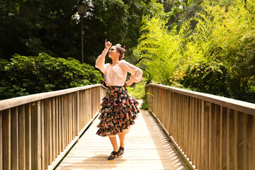 Beautiful teenage flamenco dancer dressed in a flowery skirt with ruffles and flowers in her hair is dancing in the park on a wooden bridge. Flamenco concept cultural heritage of humanity. - obrazy, fototapety, plakaty
