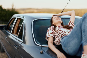 The woman driver stopped on the road and lay down on the car to rest and look at the beautiful landscape in a striped T-shirt and jeans. Complicated journey to nature