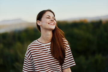 Woman traveler in nature in a striped t-shirt smiling and looking at the sunset