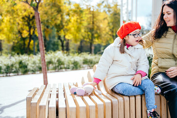Portrait of young mother who sitting on bench outdoor with cute beautiful disabled daughter and smiling, communication to each other.