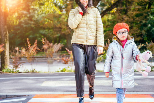 Young caucasian mother crossing street and commuting downtown city with little daughter wearing eyeglasses and red beret hold a toy in autumn time. Sun glare effect.