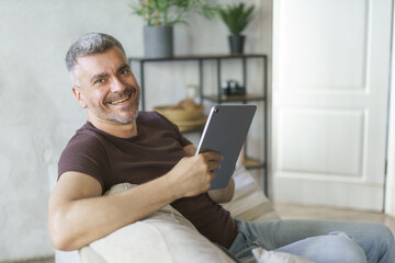 Portrait of smiling middle aged man using digital tablet pc sitting on the sofa in home interior. Grey haired freelancer working remotely at home using tablet. Business from home