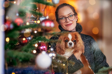 Smiling young woman holding a puppy in her hand while standing next to the christmas tree