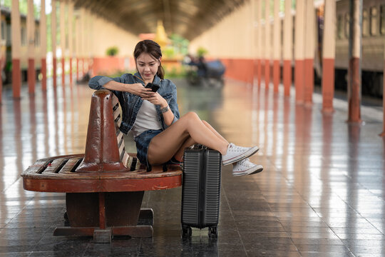 Young and beautiful woman using a phone waiting with luggage for the train at the railway station.