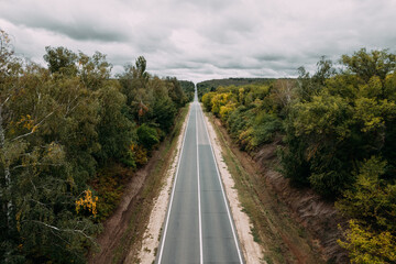Aerial top view of the empty road between green and yellow autumn trees and cloudy sky. Drone shot of an asphalt road in the countryside.