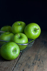 Green apples are placed in a stainless steel basket and rest on a rustic wooden floor.