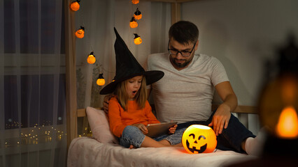 Happy family, father and little girl in witch black hat watch horror film on tablet in dark with orange pumpkin lantern at home. Halloween celebration