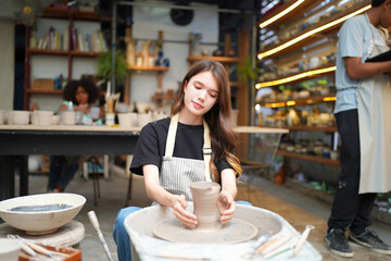 Close-up of concentrated beautiful craftswoman in apron sitting at pottery wheel and using craft tool while shaping wet clay vessel