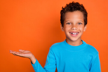 Photo portrait of cute little schoolboy palm hold empty space toothy smile wear trendy blue clothes isolated on orange color background