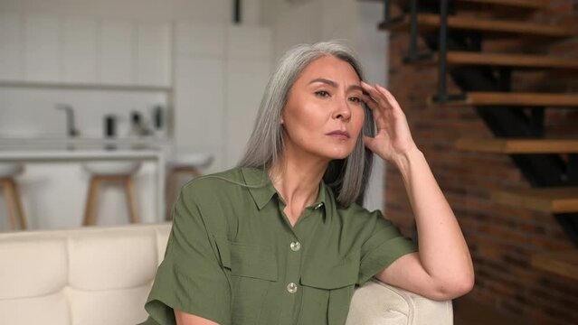 Depression concept. Upset senior gray-haired Asian woman has a feelings of severe despondency and dejection, sitting on the sofa at home, lost in thoughts and worries, pensive lady in doubt