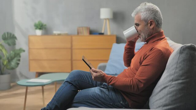 Man Sitting On Couch Watching Reading News On Tablet Device,mature Male Sits On Sofa At Home Starts The Day Looking At Day's Schedule Enjoying Coffee Cup