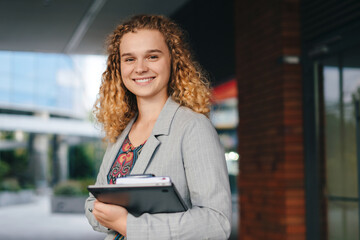 Portrait of woman university student happily holding laptop outdoors after school on campus. People...