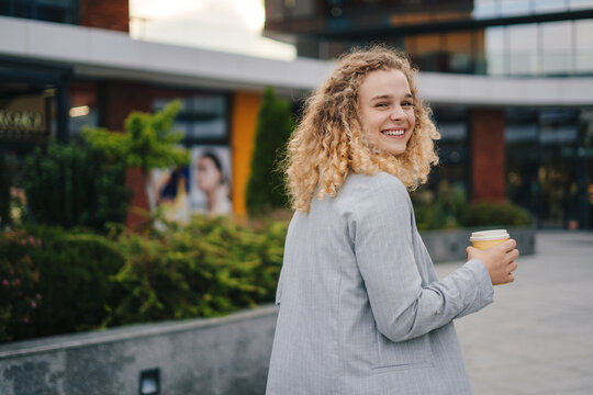 Photo of attractive lady student turning her head towards the camera while heading towards the entrance of the university, holding coffee to go and a laptop in