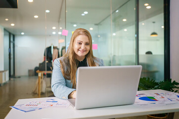 Happy female entrepreneur types on laptop computer, works in modern workspace in business center, dressed in formal outfit, surfes internet, watches training webinar, chats online with partners