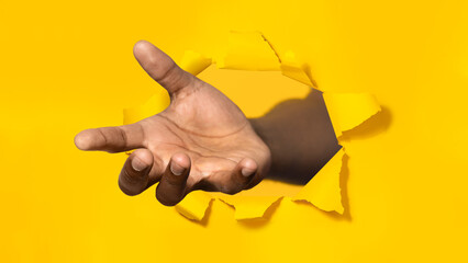African american man stretching open palm through hole in torn yellow paper, offering helping hand,...