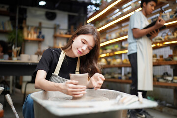 Close-up of concentrated beautiful craftswoman in apron sitting at pottery wheel and using craft tool while shaping wet clay vessel