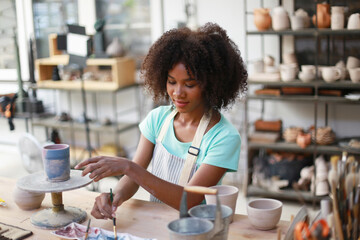 Fototapeta premium Close-up of concentrated beautiful craftswoman in apron sitting at pottery wheel and using craft tool while shaping wet clay vessel