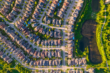 Warm summer Golden Hour evening and middle class residential houses in very geometrical setting pattern close to each other. Green grass front yards and backyards. Car roads and pedestrian walkways.