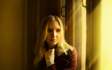 A beautiful sad blonde woman, dressed in a coat, is sitting near an old window, illuminated by rays of sunlight. Loneliness.