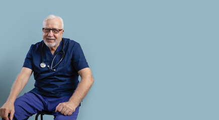 Charismatic elderly man, doctor surgeon, sitting and smiling, wearing glasses with stethoscope, in medical clothes on blue background, medicine concept, place for text, banner