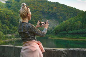 Travel memories - concept. A woman with blond hair travels in a beautiful landscape and takes photos with her smartphone. 